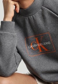 Felpa grigia con colletto alto, caratterizzata da un logo rettangolare arancione che dice "CALVIN KLEIN JEANS" in carattere bold. Texture morbida.