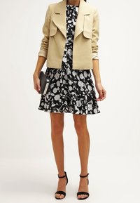 Beige kort jacka över en svart klänning med vitt blommönster. Svarta sandaletter med remmar och en liten handväska. Släta texturer.