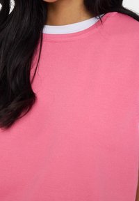 Kvinna med långt mörkt hår som bär en rosa tröja över en vit skjorta, vilket visar halslinning och överkropp.