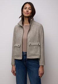 Graue Fischgrätenjacke mit hohem Kragen, zwei Fronttaschen und Reißverschluss. Kombiniert mit einem hellbraunen Oberteil und blauen Jeans.