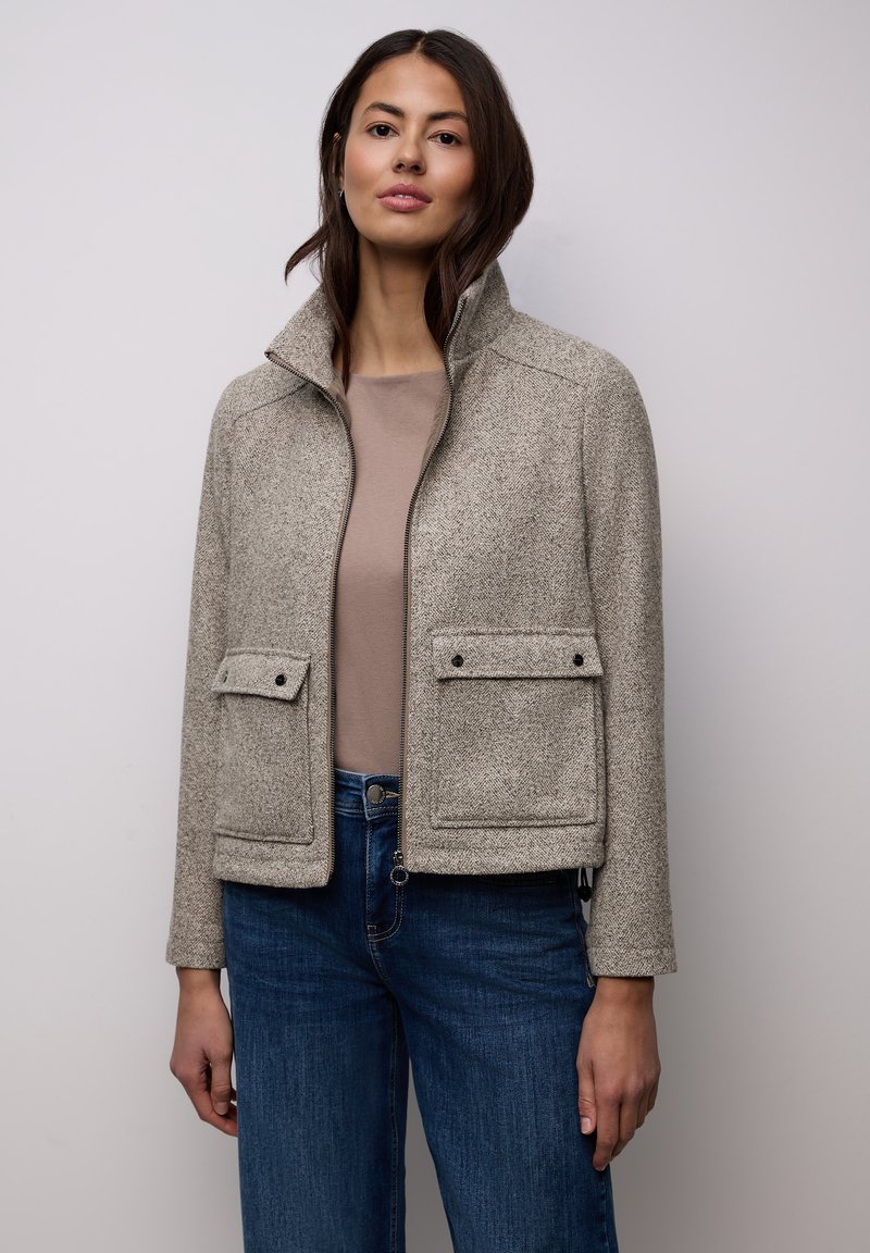 Graue Fischgrätenjacke mit hohem Kragen, zwei Fronttaschen und Reißverschluss. Kombiniert mit einem hellbraunen Oberteil und blauen Jeans.