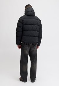 Chaqueta acolchada negra con capucha, con secciones acolchadas, llevada sobre una camisa de cuadros, combinada con jeans oscuros y zapatos marrones.