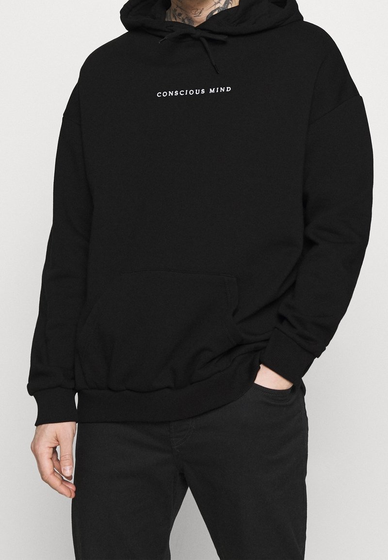 Sudadera negra con un bolsillo frontal, con el texto "MENTE CONSCIENTE" en blanco. Confeccionada con una tela suave y un corte relajado.