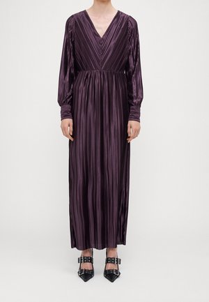 Kobieta w długiej, plisowanej sukni maxi w głębokim odcieniu purpury z dekoltem w serek, nosząca czarne szpilki z klamrą na czubku, na jednolitym tle.