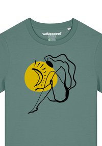 Groene katoenen t-shirt met een zwarte lijntekening van een figuur naast een gele cirkel met een zon- en maanontwerp.