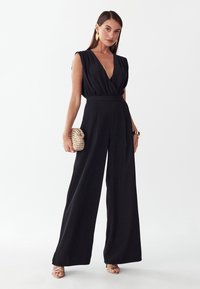 Frau trägt einen ärmellosen schwarzen Jumpsuit mit weitem Bein und tiefem V-Ausschnitt, hält eine strukturierte goldene Clutch und trägt goldene Sandalen und Ohrringe.