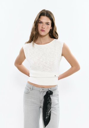 Junge Frau mit langem braunem Haar, die ein ärmelloses weißes Strickoberteil und hellgraue Jeans trägt, an deren Gürtelschlaufe ein schwarzer Schal mit Muster gebunden ist.