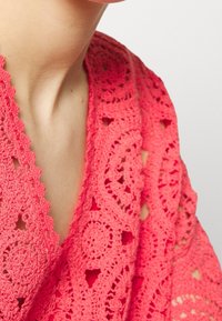 Top de crochet en color coral con intrincados patrones circulares y bordes festoneados, que presenta un tejido abierto texturizado y un diseño de escote en V.