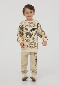 Felpa beige con grafica di un orso e testo colorato, abbinata a pantaloni jogger beige con un design a contrasto. Tessuto morbido, vestibilità comoda.