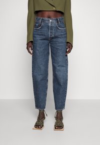 Jeans in denim blu a vita alta con gamba dritta, dotati di cinque tasche e cuciture a contrasto. Indossati con sandali neri e dorati con cinturini.