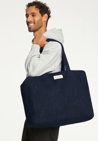 Sac fourre-tout bleu marine avec un extérieur texturé, des poignées solides et une forme rectangulaire. Présente une petite étiquette à l'avant.