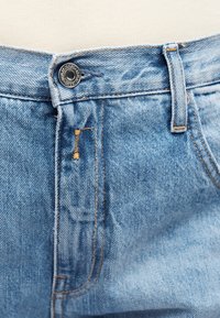 Κοντινό πλάνο της ζώνης μέσης του ανοιχτόμπλε τζιν με ασημένιο κουμπί και εμφανείς ραφές.