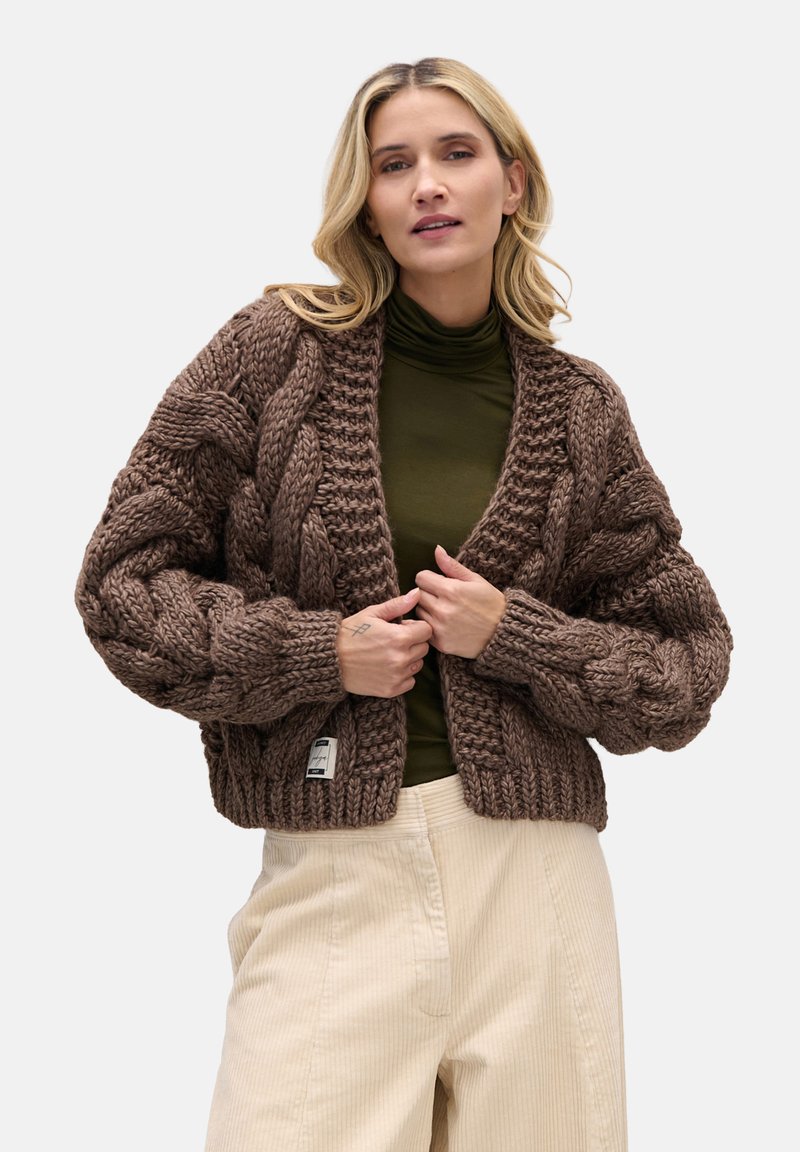 Brązowy, gruby sweter na guziki z fakturowanymi warkoczami, krótki krój, z otwartym przodem, noszony na ciemnej bluzce i zestawiony z jasnymi spodniami sztruksowymi.