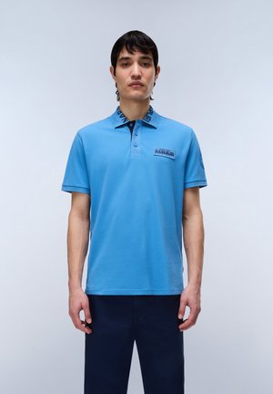 Mand iført en lyseblå polotrøje med mørkeblå tekst på kraven og patch-detalje, kombineret med mørkeblå bukser, stående mod en ensfarvet baggrund.