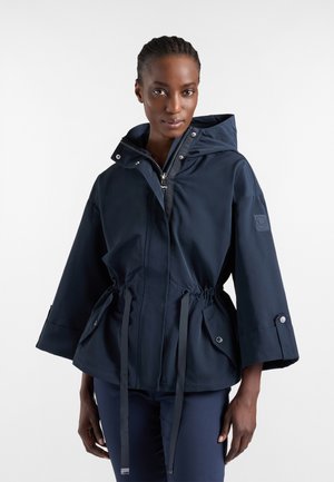 Donna con capelli intrecciati indossa una giacca blu navy con cappuccio, vita con coulisse, chiusure con zip e bottoni a pressione e grandi tasche frontali.