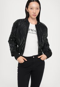 Giubbino bomber nero con una superficie lucida a onde, polsini a coste e chiusura con zip; abbinato a una t-shirt bianca e jeans neri.