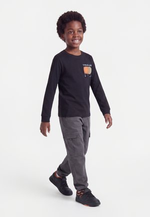 Garçon souriant qui marche, portant un t-shirt noir à manches longues avec un motif citrouille, un pantalon cargo gris et des baskets noires avec des détails orange.