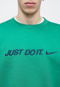 Πράσινο φούτερ με στρογγυλή λαιμόκοψη, με κεντητό κείμενο "JUST DO IT." σε σκούρο μπλε και λογότυπο Nike δεξιά. Μαλακό υλικό.