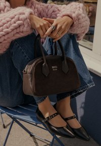 Borsa sintetica marrone con due manici e logo dorato. Ballerine nere con cinturino alla caviglia. Modella che indossa un maglione rosa a maglia e jeans blu.