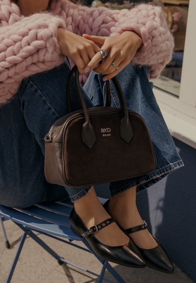 Borsa sintetica marrone con due manici e logo dorato. Ballerine nere con cinturino alla caviglia. Modella che indossa un maglione rosa a maglia e jeans blu.