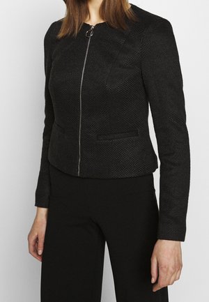 Frau trägt eine enganliegende schwarze, strukturierte Reißverschlussjacke mit langen Ärmeln, kombiniert mit schwarzen Hosen, vor einem schlichten hellen Hintergrund.