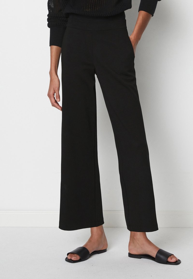 Pantalon noir à jambes larges en tissu doux et extensible avec une taille haute. Associé à des chaussures plates noires sans lacets ; design et couleur minimalistes.