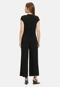 Zwarte jumpsuit met kapmouwen, wijde broekspijpen, gladde stof en een getailleerde taille. Draagt een paar zwarte wedge sandalen, gezien vanuit de achterkant.