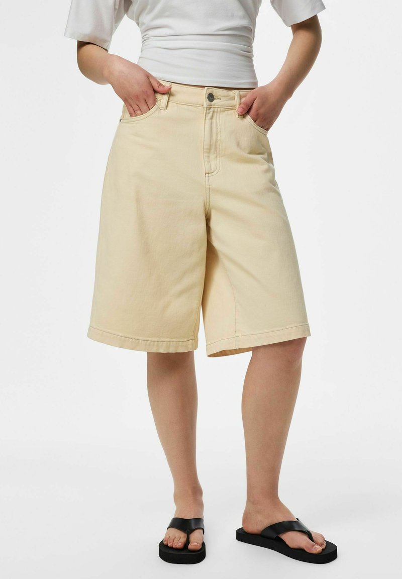 Personne portant un short beige au niveau des genoux, un haut blanc à manches courtes et des tongs plateformes noires, les mains dans les poches du short.