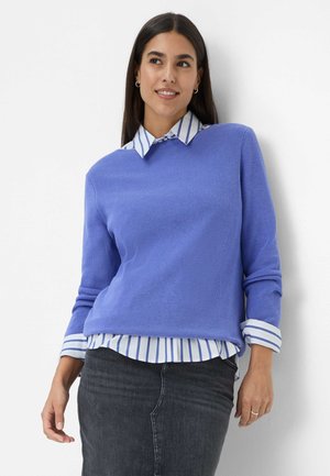 Lichtblauwe geribde trui over een wit shirt met blauwe strepen. Heeft lange mouwen en een ronde halslijn, gecombineerd met donkerdenim.