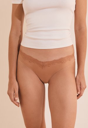 Femme portant des sous-vêtements beiges sans coutures avec bordure en dentelle et un t-shirt blanc court, debout devant un fond beige neutre.