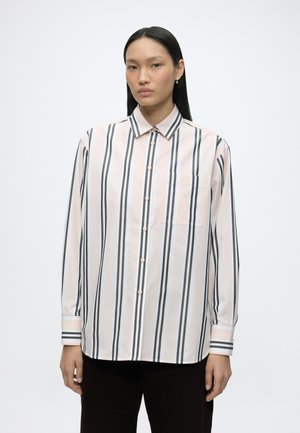 Vrouw met lang zwart haar die een lichtroze en zwarte verticaal gestreepte blouse met knopen en een zwarte broek draagt tegen een witte achtergrond.