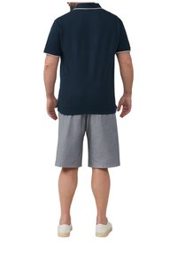 Marineblaues Poloshirt mit heller Akzentuierung, kombiniert mit grauen gemusterten Shorts. Das Modell trägt weiße Sneakers; die Ansicht ist von hinten.