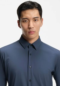 Jonge man met kort donker haar, gekleed in een keurig dichtgeknoopt donkerblauw overhemd met kraag, die recht vooruit kijkt tegen een eenvoudige lichte achtergrond.