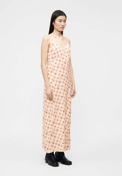 Γυναίκα που στέκεται, φορώντας ένα μακρύ, ροζ ανοιχτό φόρεμα με λεπτές τιράντες και μαύρα μποτάκια στους αστραγάλους, σε απλό λευκό φόντο.