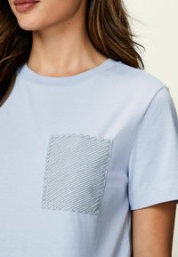 Vaaleansinen puuvillapaita, jossa on teksturoitu tasku, jossa on siksak-kuvio ommeltuna. Pyöreä kaula-aukko ja lyhyet hihat viimeistelevät suunnittelun.