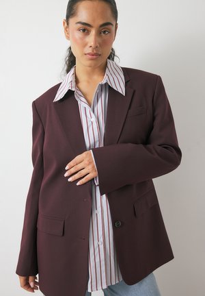 Burgundy blazer with structured shoulders, single button closure, and two front pockets, layered over a white striped shirt with button details.