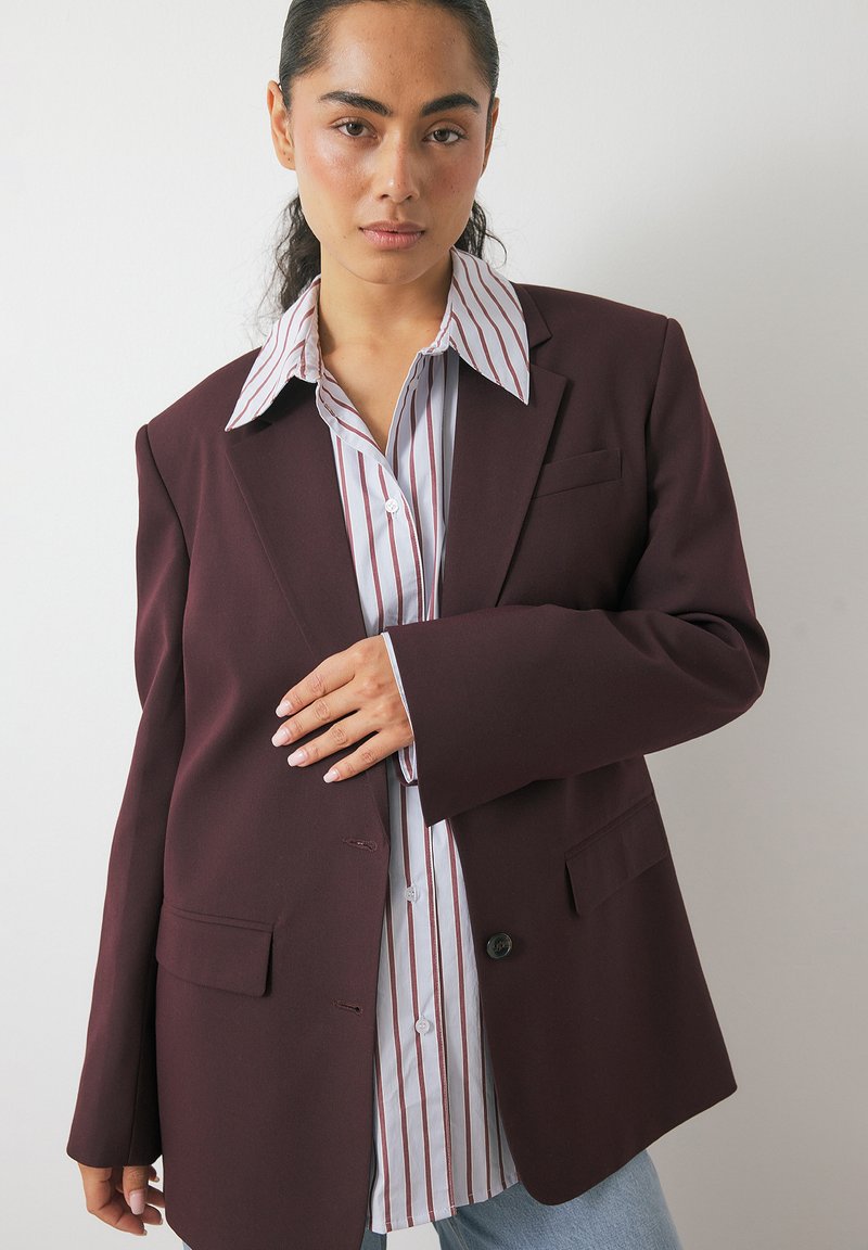 Bordeaux blazer met gestructureerde schouders, sluiting met één knop en twee voorzakken, gedragen over een wit gestreept shirt met knoopdetails.