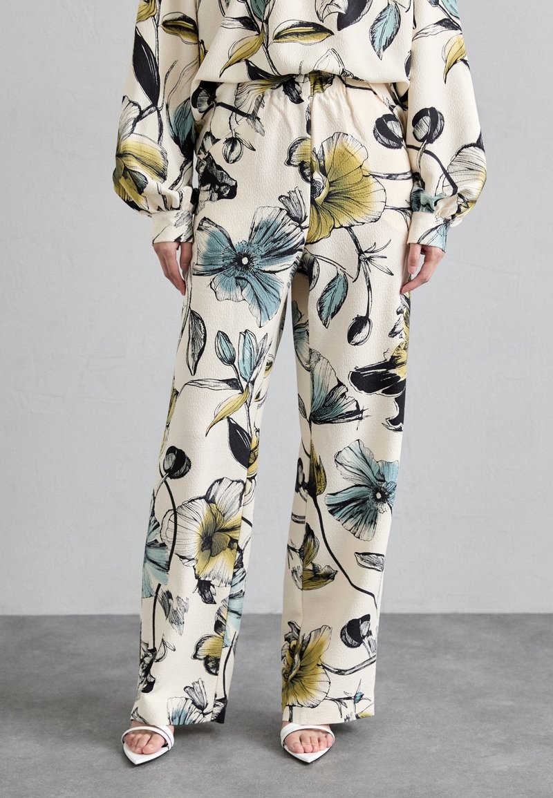 Pantalon crème à jambes larges avec un grand imprimé floral bleu, jaune et noir, porté avec des talons blancs à bouts ouverts sur un sol gris.