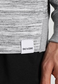 Suéter gris jaspeado con textura acanalada y una etiqueta blanca con la inscripción "ONLY & SONS" en el lado inferior izquierdo, usado sobre pantalones negros.