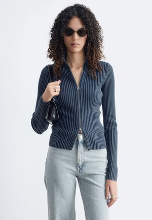 Femme aux cheveux bouclés portant des lunettes de soleil, un pull zippé côtelé sombre, un jean bleu clair, et tenant un petit sac à main noir sur l’épaule.