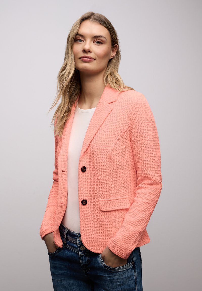 Jeune femme aux longs cheveux blonds portant un blazer corail texturé sur une chemise blanche et un jean bleu, les mains dans les poches, sur un fond uni.
