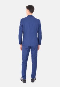 Completo blu realizzato in tessuto liscio, vestibilità su misura, con una sola fessura sul retro. Abbinato a pantaloni coordinati e scarpe da dress nere.
