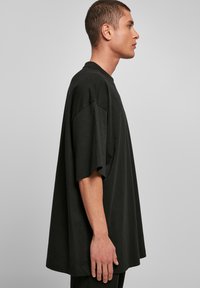 Zwarte oversized t-shirt met een hoge halslijn en korte mouwen. Zachte textuur, losvallende pasvorm en rechte zoom. Model wordt vanaf de zijkant getoond.