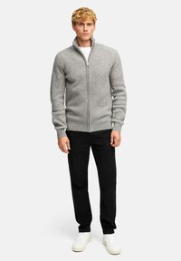 Szary sweter z prążkowanej dzianiny z wysokim kołnierzem i zamkiem błyskawicznym, połączony z czarnymi spodniami i białymi trampkami. Tło w naturalnym świetle.