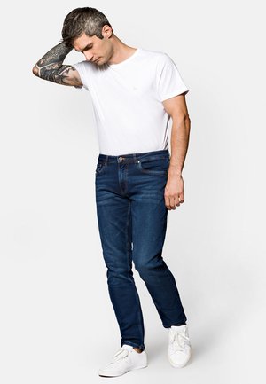 Ciemnoniebieskie dopasowane dżinsy z lekkim przetarciem, połączone z prostą białą koszulką. Stylizację uzupełniają białe trampki.