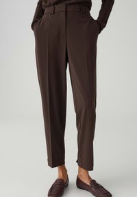 Bruine op maat gemaakte broek tot aan de enkels, gedragen met bijpassende bruine loafers en handen in de zakken tegen een effen achtergrond.