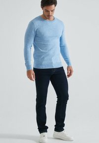 Pull bleu clair à manches longues avec un col rond et des poignets côtelés, associé à un jean foncé et des chaussures de sport blanches.
