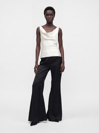 Femme debout portant un top blanc sans manches à col bénitier et un pantalon noir à jambes larges sur un fond gris uni.