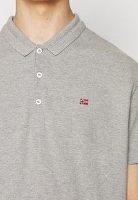 Personne portant un polo gris clair avec trois boutons blancs et un petit drapeau norvégien brodé sur la poitrine.