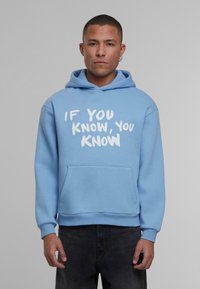 Svetlo modra kapuca iz mehkega blaga, z sprednjo žepno in belim napisom "ČE VEŠ, VEŠ" v drznem slogu čopiča.